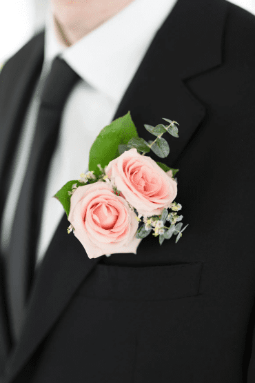 groom wearing a black suite and a boutonniere with 2 blue pink roses and greenery for wedding