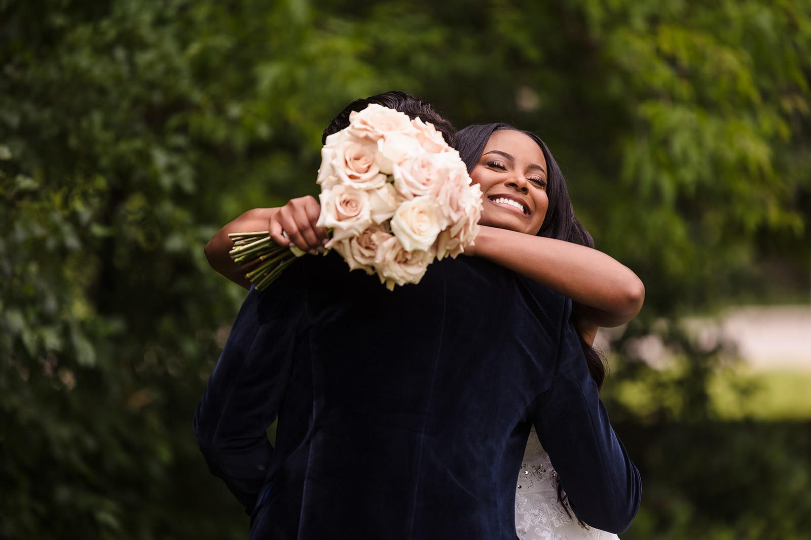 A bride and groom share an embrace in a lush green outdoor setting. The groom, facing away from the camera, holds a bouquet of white and pink roses. The bride, facing the camera and smiling broadly, leans into the hug, showing joy and happiness.