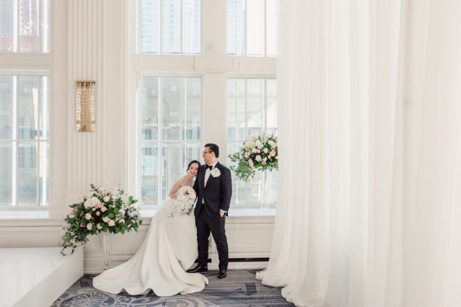 Bride and groom standing in front of a window at The Omni king Edward hotel wedding venue in Toronto, with 2 large floral arrangements on either side of them. one on the ground and one by the window ledge. Done with white, blush pink and accents of cappuccino tones and greenery with some cascading parts, sitting on glass vases.