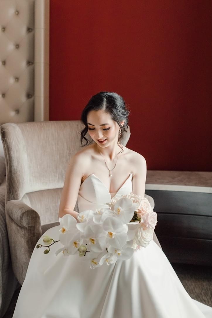 Bride sitting on a chair holding her beautiful bridal bouquet of cascading white orchids and white and dusty pink roses.